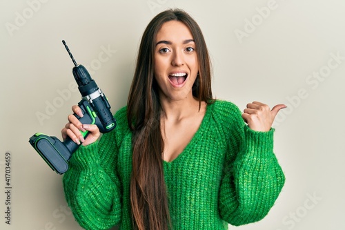 Young hispanic girl holding screwdriver pointing thumb up to the side smiling happy with open mouth
