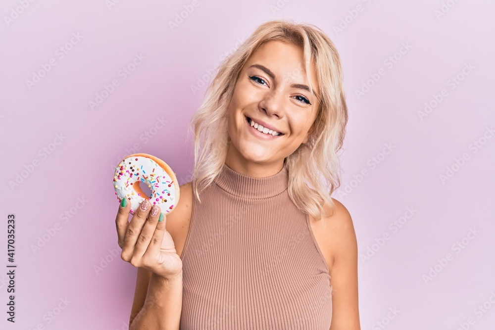 Young blonde girl holding donut looking positive and happy standing and smiling with a confident smile showing teeth Young blonde girl holding donut looking positive and happy standing and smiling with a confident smile showing teeth