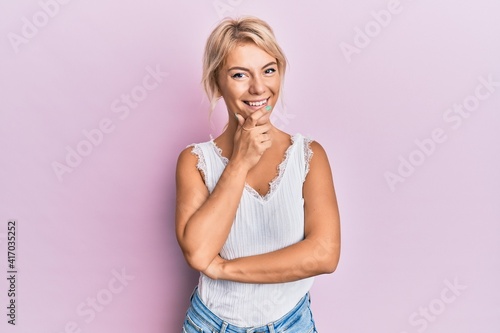 Young blonde girl wearing casual clothes looking confident at the camera with smile with crossed arms and hand raised on chin. thinking positive.