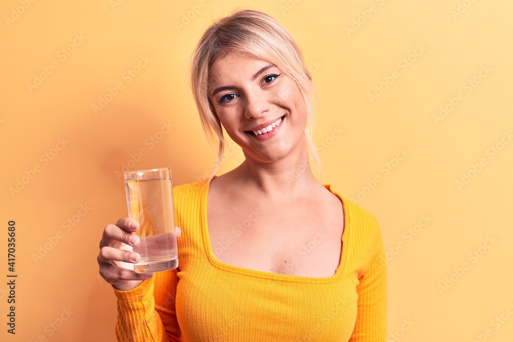 Young beautiful blonde woman drinking glass of water over isolated yellow background looking positive and happy standing and smiling with a confident smile showing teeth Young beautiful blonde woman drinking glass of water over isolated yellow background looking positive and happy standing and smiling with a confident smile showing teeth