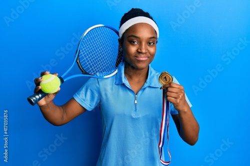 African american woman with braided hair playing tennis holding racket and ball and winner medal relaxed with serious expression on face. simple and natural looking at the camera. © Krakenimages.com