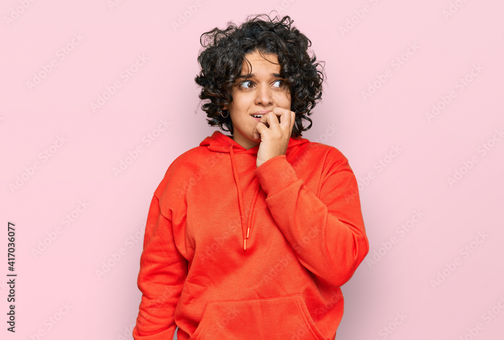 Young hispanic woman with curly hair wearing casual sweatshirt looking stressed and nervous with hands on mouth biting nails. anxiety problem. Young hispanic woman with curly hair wearing casual sweatshirt looking stressed and nervous with hands on mouth biting nails. anxiety problem.