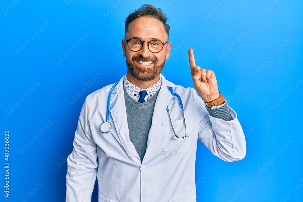 Handsome middle age man wearing doctor uniform and stethoscope showing and pointing up with finger number one while smiling confident and happy. Handsome middle age man wearing doctor uniform and stethoscope showing and pointing up with finger number one while smiling confident and happy.