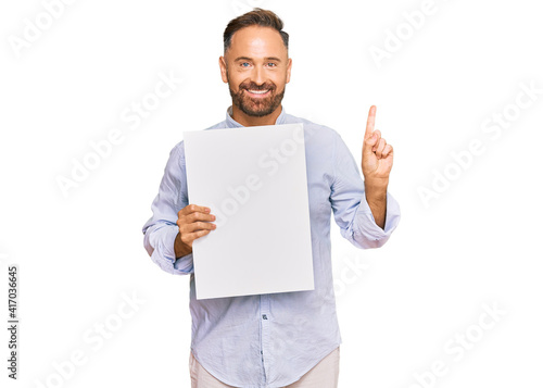 Handsome middle age man holding blank empty banner surprised with an idea or question pointing finger with happy face, number one