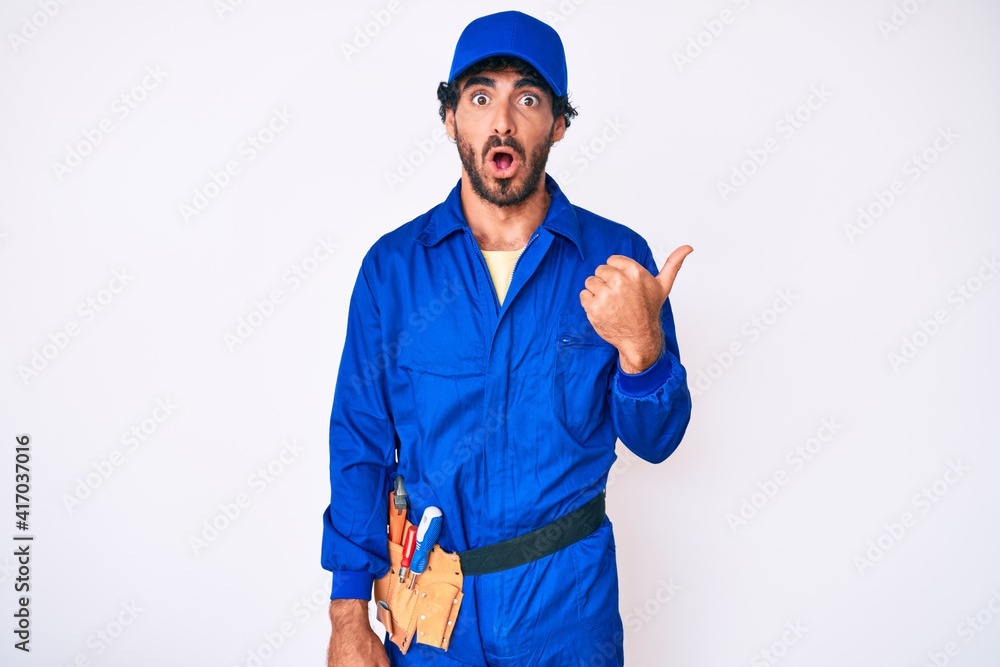 Handsome young man with curly hair and bear weaing handyman uniform surprised pointing with hand finger to the side, open mouth amazed expression.