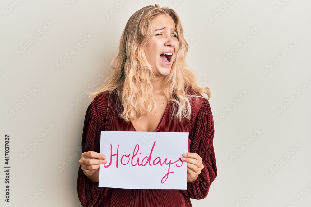 Young blonde girl holding holidays banner message angry and mad screaming frustrated and furious, shouting with anger. rage and aggressive concept. Young blonde girl holding holidays banner message angry and mad screaming frustrated and furious, shouting with anger. rage and aggressive concept.
