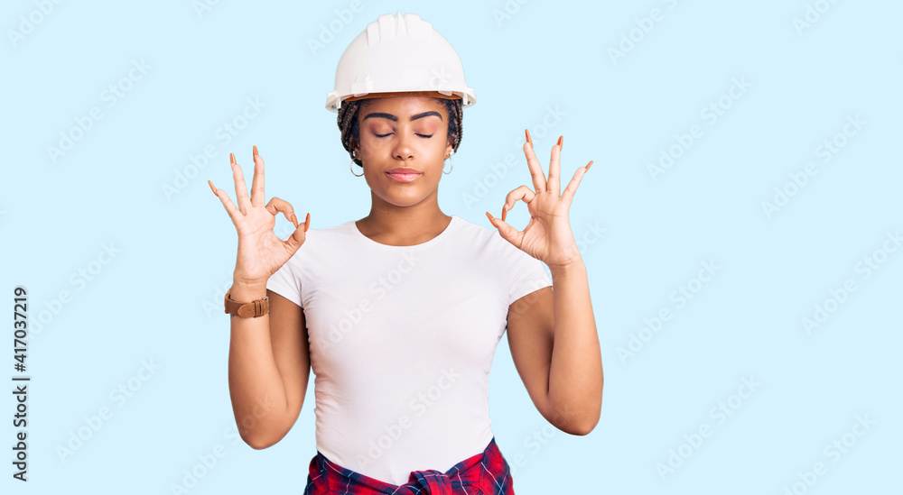 Young african american woman with braids wearing hardhat and builder clothes relax and smiling with eyes closed doing meditation gesture with fingers. yoga concept. Young african american woman with braids wearing hardhat and builder clothes relax and smiling with eyes closed doing meditation gesture with fingers. yoga concept.