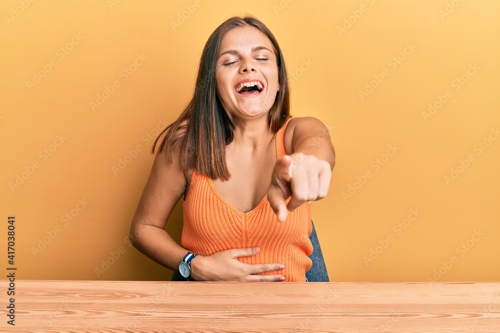 Young caucasian woman wearing casual clothes sitting on the table laughing at you, pointing finger to the camera with hand over body, shame expression Young caucasian woman wearing casual clothes sitting on the table laughing at you, pointing finger to the camera with hand over body, shame expression