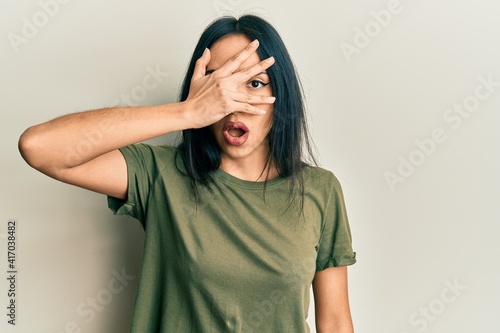 Young hispanic girl wearing casual t shirt peeking in shock covering face and eyes with hand, looking through fingers with embarrassed expression. © Krakenimages.com