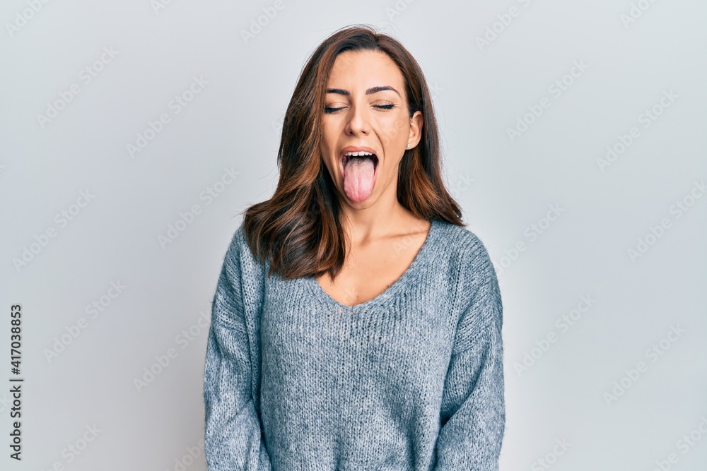 Young brunette woman wearing casual winter sweater sticking tongue out happy with funny expression. emotion concept. - obrazy, fototapety, plakaty 