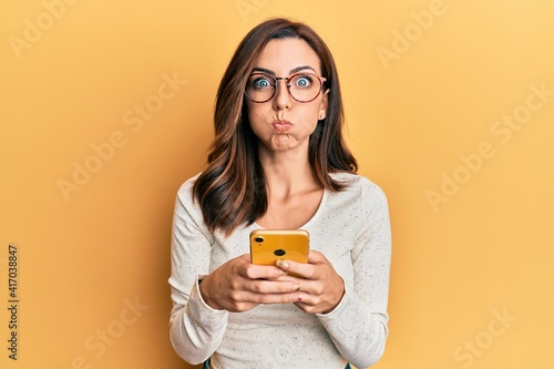 Tableau sur toile Young brunette woman using smartphone over yellow background puffing cheeks with funny face