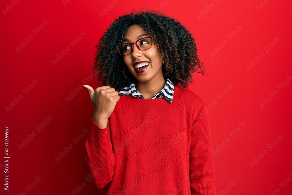 Beautiful african american woman with afro hair wearing sweater and glasses smiling with happy face looking and pointing to the side with thumb up. Beautiful african american woman with afro hair wearing sweater and glasses smiling with happy face looking and pointing to the side with thumb up.