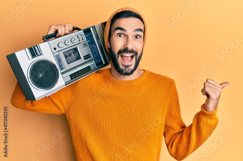 Young hispanic man holding boombox, listening to music pointing thumb up to the side smiling happy with open mouth