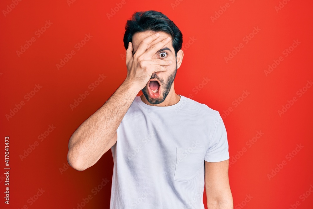 Young hispanic man wearing casual white tshirt peeking in shock covering face and eyes with hand, looking through fingers with embarrassed expression. Young hispanic man wearing casual white tshirt peeking in shock covering face and eyes with hand, looking through fingers with embarrassed expression.