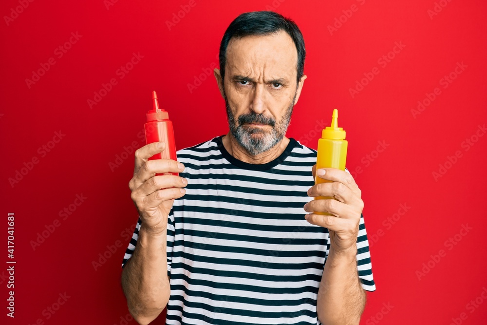 Middle age hispanic man holding ketchup and mustard bottle skeptic and nervous, frowning upset because of problem. negative person. Middle age hispanic man holding ketchup and mustard bottle skeptic and nervous, frowning upset because of problem. negative person.