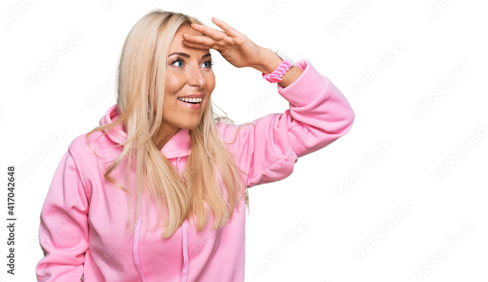 Young blonde woman wearing casual sweatshirt very happy and smiling looking far away with hand over head. searching concept. Young blonde woman wearing casual sweatshirt very happy and smiling looking far away with hand over head. searching concept.