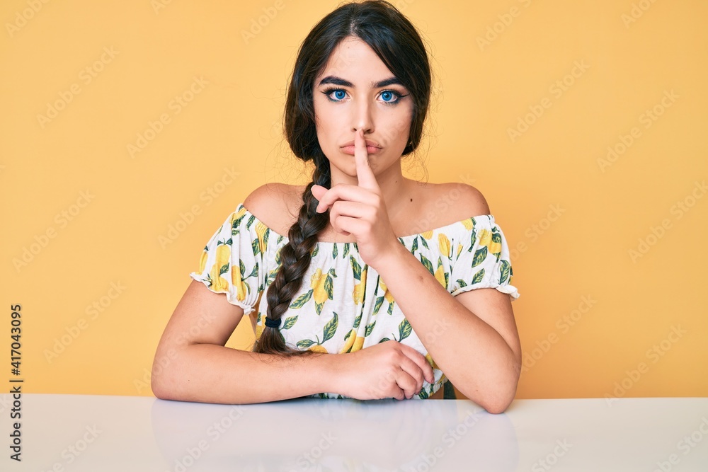 Brunette teenager girl wearing casual clothes sitting on the table asking to be quiet with finger on lips. silence and secret concept. Brunette teenager girl wearing casual clothes sitting on the table asking to be quiet with finger on lips. silence and secret concept.
