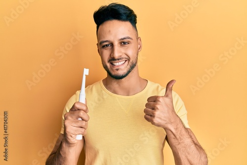 Young arab man holding electric toothbrush smiling happy and positive, thumb up doing excellent and approval sign