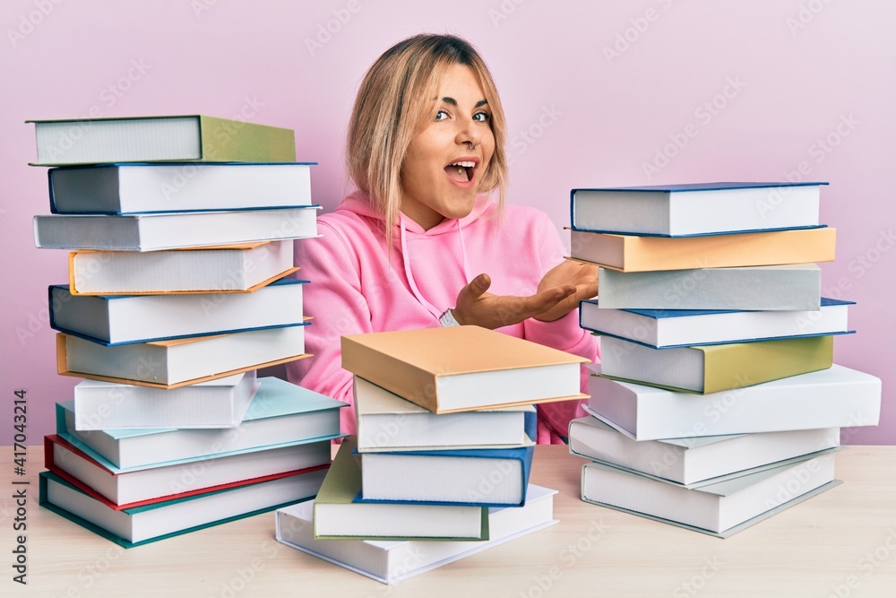 Young caucasian woman sitting on the table with books pointing aside with hands open palms showing copy space, presenting advertisement smiling excited happy Young caucasian woman sitting on the table with books pointing aside with hands open palms showing copy space, presenting advertisement smiling excited happy