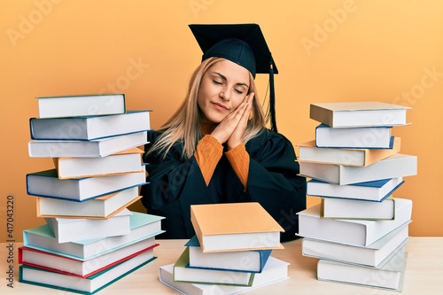 Young caucasian woman wearing graduation ceremony robe sitting on the table sleeping tired dreaming and posing with hands together while smiling with closed eyes.