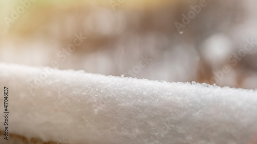 A thick layer of snow on the background of trees. Winter holidays background with snow texture. Layer of snow on a fence whit trees in the background. Sunny day, rays glow. Snowflakes falling. Shine