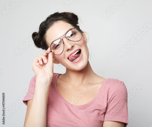 Younf brunette woman wearing pink thirt and glasses showing tongue and looking at the camera over grey background