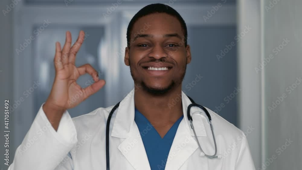 Positive african american male doctor showing okay gesture and smiling widely at camera, clinic interior, slow motion Positive african american male doctor showing okay gesture and smiling widely at camera, clinic interior, slow motion