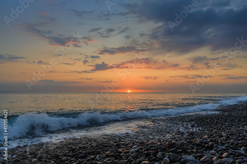 Sunset over the sea. In the foreground  a wave running onto a pebbly shore with white foam. The dark blue sky with clouds  on the horizon line the orange colors of the sunset and the circle of the sun