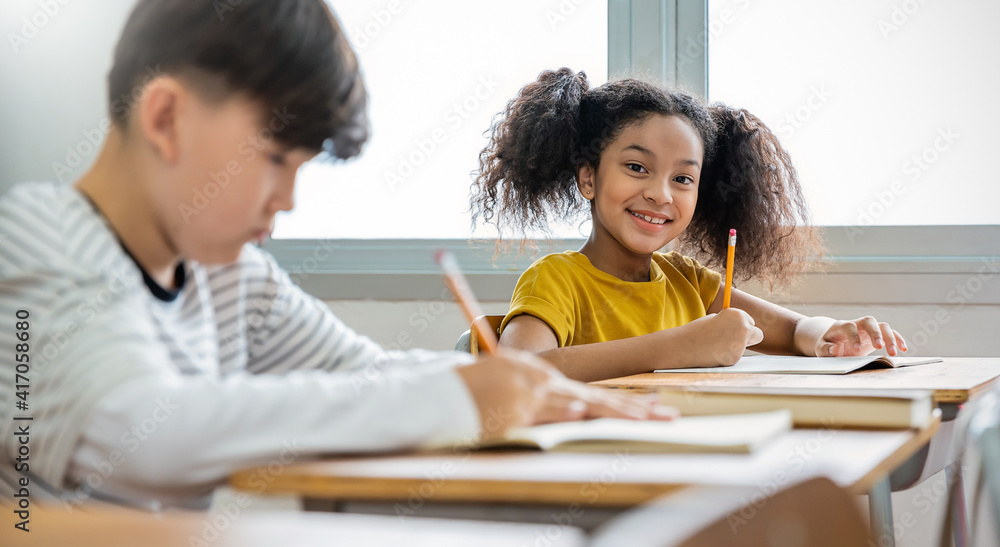 Portrait of little pupil writing at desk in classroom at the elementary school. Student African girl doing test in primary school. Children writing notes in classroom. Education knowledge banner Portrait of little pupil writing at desk in classroom at the elementary school. Student African girl doing test in primary school. Children writing notes in classroom. Education knowledge banner