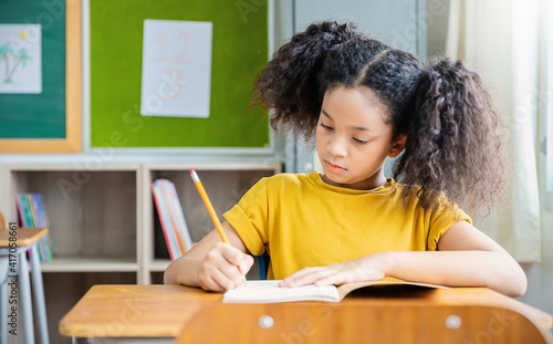 Portrait of little pupil writing at desk in classroom at the elementary school. Student African girl doing test in primary school. Children writing notes in classroom. Education knowledge (blur)
