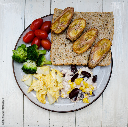 Top down view of Healthy Breakfast dish on a white wooden background, Bread with baked bananas, scrambled eggs with onions, red beans, cherry tomatoes and broccoli on a dish