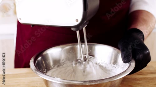 chef in black gloves hammer egg white in a metal bowl on a wooden table using an electric mixer