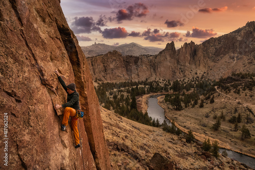 Adventurous man is rock climbing on the side of a steep cliff. Sunset Sky Art Render. Taken in Smith Rock, Oregon, North America.