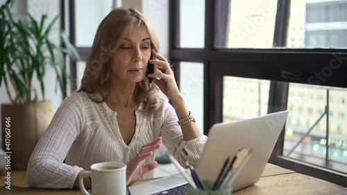 Businesswoman having mobile call in front of laptop screen at table in home office. Middle aged woman talking about something and looking at computer monitor, sitting at desk in light room, coffee mug