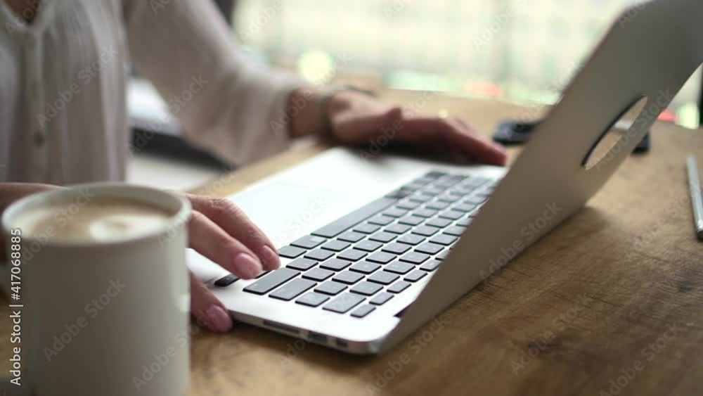 Businesswoman typing on laptop keyboard and sitting at table in home office. Closeup view of middle aged American woman types text in front of computer monitor and sits at wooden desk in light room Businesswoman typing on laptop keyboard and sitting at table in home office. Closeup view of middle aged American woman types text in front of computer monitor and sits at wooden desk in light room