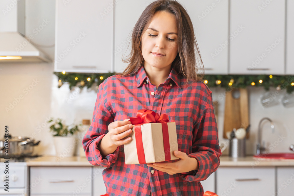 Young woman in a red shirt holding box with red ribbon in the kitchen for the holiday. Girl looks gift and smiles, becomes happy Young woman in a red shirt holding box with red ribbon in the kitchen for the holiday. Girl looks gift and smiles, becomes happy