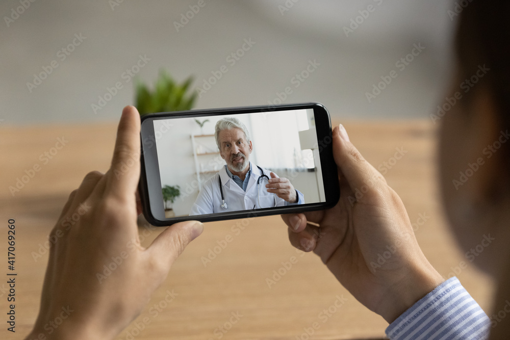 Back view close up of woman look at cellphone screen have online consultation with make mature doctor. Female speak talk on video call on smartphone with elderly GP, consult about health problem.