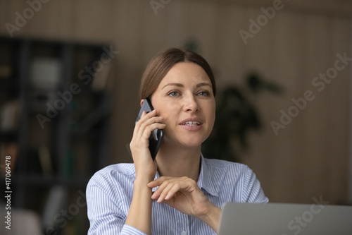 Young Caucasian woman work on computer in office talk on cellphone with client customer. Millennial female speak on smartphone at home use laptop, have virtual distant consultation with partner.