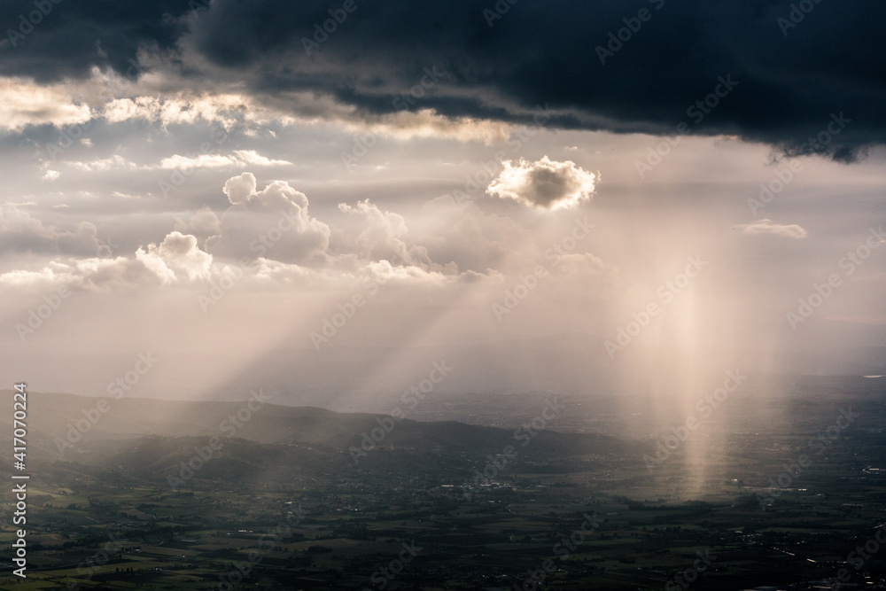 Rain, mist and sunrays between valley and layers of mountains and hills beneath a moody, overcast sky with heavy clouds