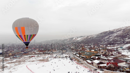 Aerial view of a hot air balloon over the icy  lake, snowy mountains around. hot air balloon in winter. Children are sledding. Glides over the camera in winter. People an ice slide.  photo