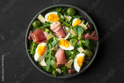 Salad with prosciutto, olives, cheese, eggs and arugula in a plate on black background. Italian food snacks. Top view