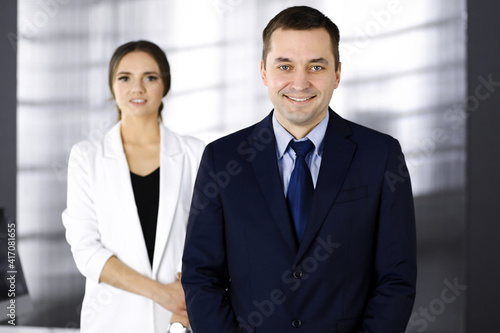 Portrait of a middle aged businessman in a dark blue suit, standing in an office with a colleague. Concept of success in a business