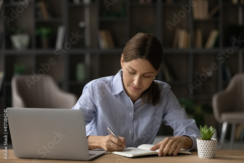 Focused millennial Caucasian woman sit at desk study online on computer writing making notes. Concentrated young female take distant course or training using laptop. Education concept.