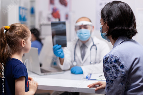 Caucasian doctor examining x-ray bones image of child and explaing treatment to prevent illness. Pediatrician specialist with protection mask and visor against covid19 providing healthcare service