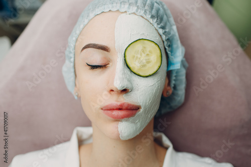 Young woman applying mask of clay on face with cucumber on eyes in beauty spa