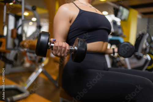 Fit woman trains biceps with dumbbells in her hands while sitting on bench in modern gym