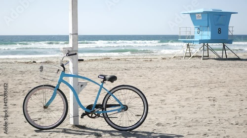 Blue bicycle, cruiser bike by ocean beach, pacific coast, Oceanside California USA. Summertime vacations, sea shore. Vintage cycle on sand near lifeguard tower or watchtower hut. Sky and water waves.