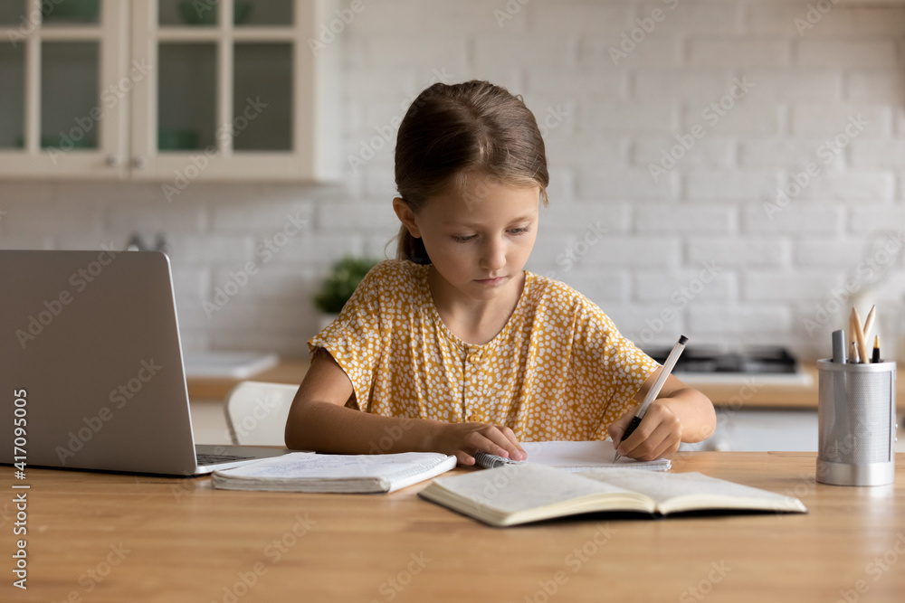 Homeschooling. Focused little schoolgirl sit at desk by laptop doing homework handwriting taking notes to paper copybook. Small junior girl engaged in distant learning get remote elementary education Homeschooling. Focused little schoolgirl sit at desk by laptop doing homework handwriting taking notes to paper copybook. Small junior girl engaged in distant learning get remote elementary education