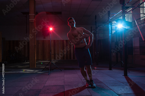 Muscular man works out with the trx system in a dark gym. Athlete with naked torso workout with resistance straps in red-blue neon light. Functional, cross training