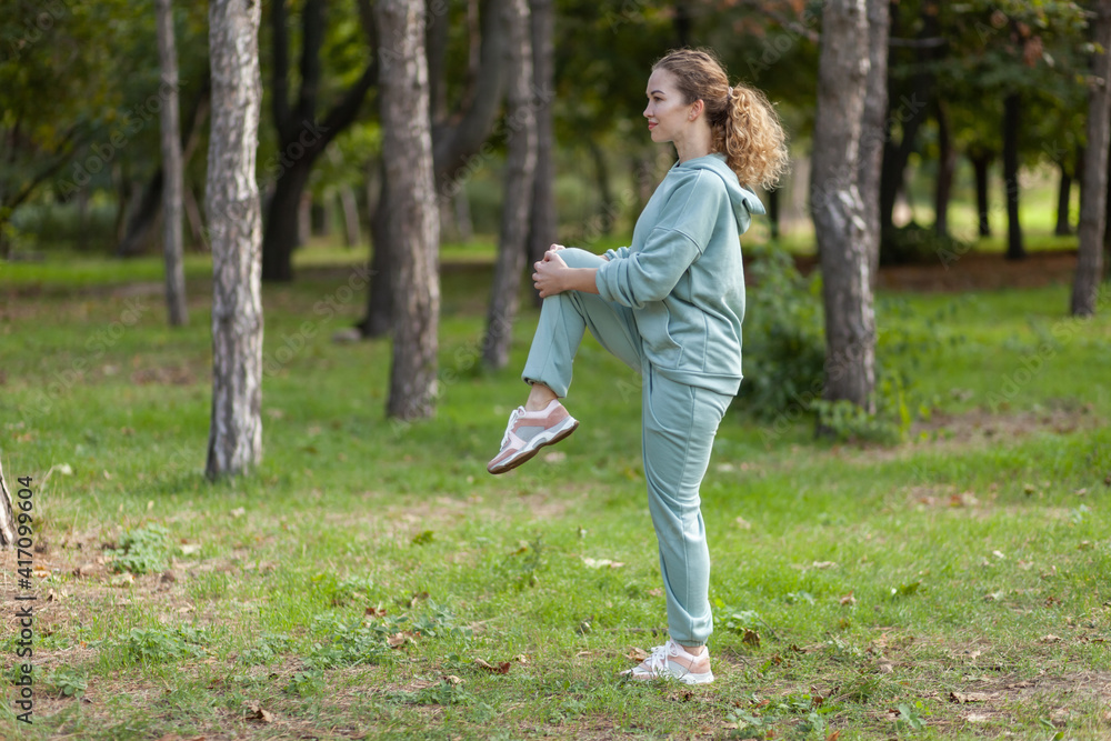 Attractive fit slim Caucasian woman with curly hair in sportswear is doing stretching exercise in the park or forest on a bright sunny day. Healthy lifestyle